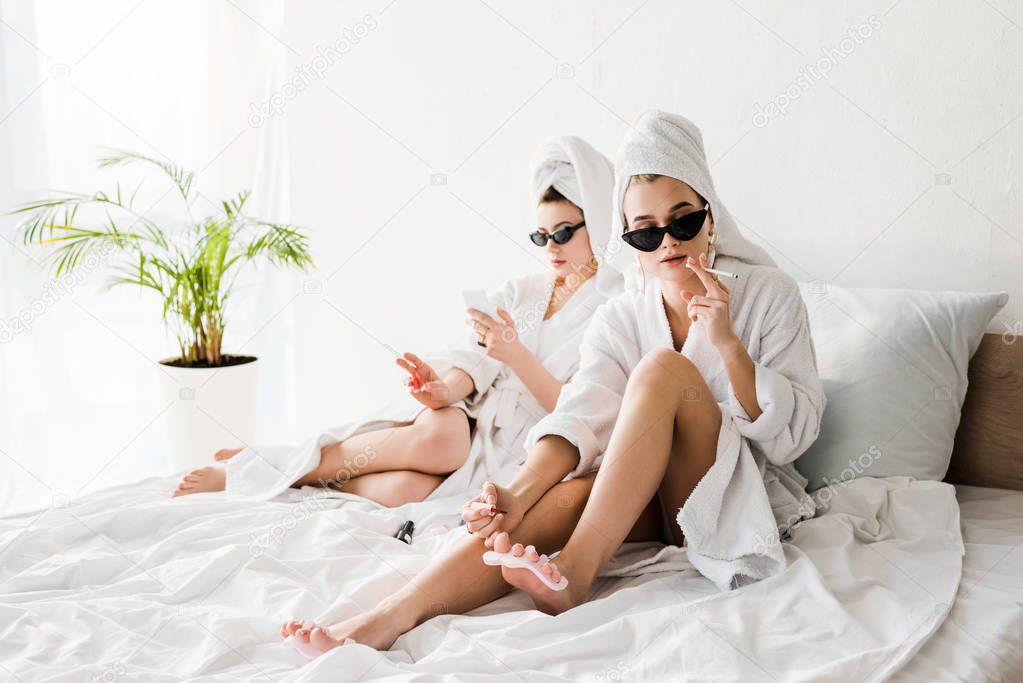 stylish women in bathrobes and sunglasses, towels and jewelry lying in bed, doing pedicure, smoking and using smartphone