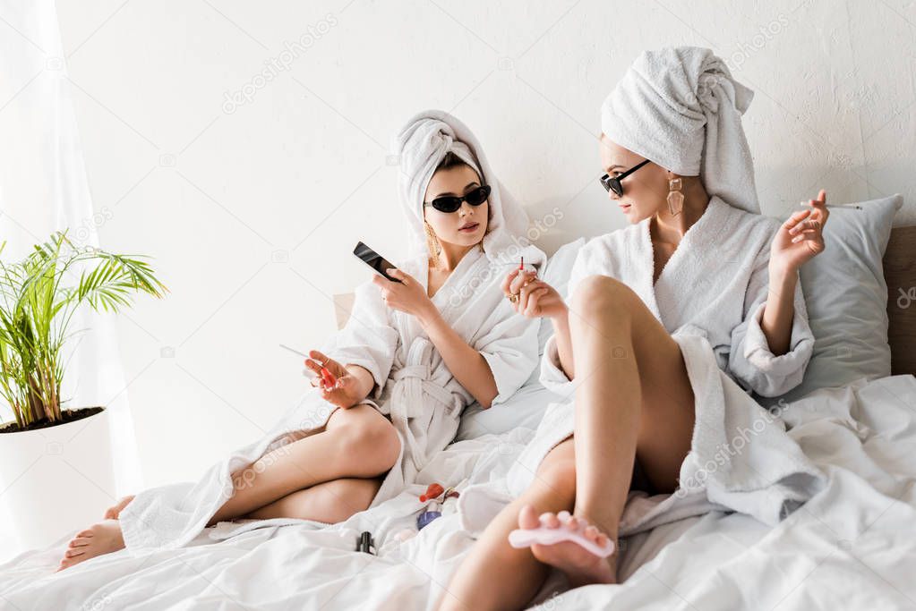 stylish women in bathrobes and sunglasses, towels and jewelry lying in bed, doing pedicure and smoking