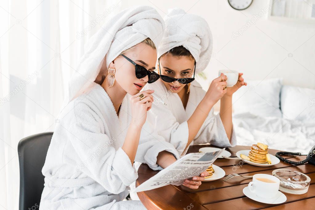 stylish women in bathrobes, sunglasses and jewelry with towels on heads smoking and reading newspaper while having breakfast 