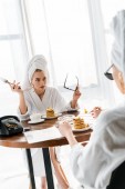 shocked stylish woman in bathrobe and jewelry with towel on head holding fork and sunglasses and staring at friend during breakfast