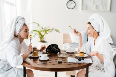 stylish happy women in bathrobes and jewelry with towels on heads talking during breakfast