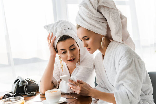 stylish women in bathrobes and jewelry with towels on heads using smartphone