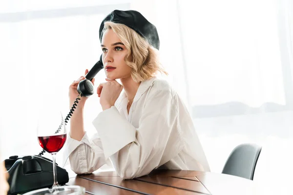 黒いベレーと白いシャツを着たエレガントなブロンドの女性がテーブルに座り テーブルの上に赤ワインとガラスの近くでOレトロな電話を話す — ストック写真
