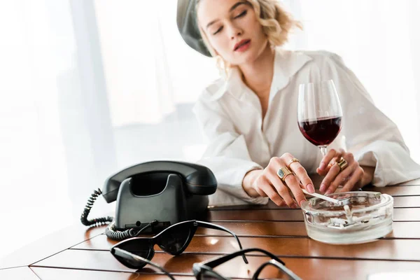 选择焦点优雅的金发女郎在黑色贝雷乌吸烟和拿着玻璃与红酒在桌子上与复古手机 — 图库照片