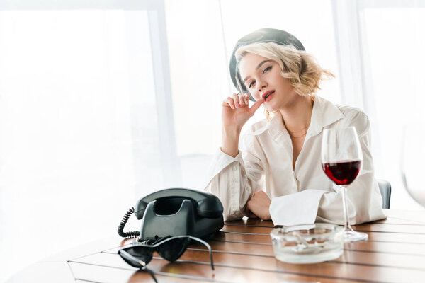 elegant blonde woman in black beret smoking cigarette and sitting at table with retro phone and glass of red wine