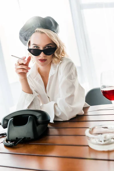 レトロな電話と赤ワインのグラスの近くに座っている間 黒いベレーとサングラスでタバコを吸うエレガントなブロンドの女性 — ストック写真