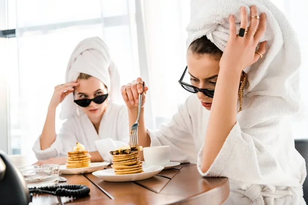 Stylish tired women in bathrobes, sunglasses and jewelry with towels on heads eating pancakes for breakfast — Stock Image Stylish Tired Women Bathrobes Sunglasses Jewelry Towels Heads Eating Pancakes Stock Picture