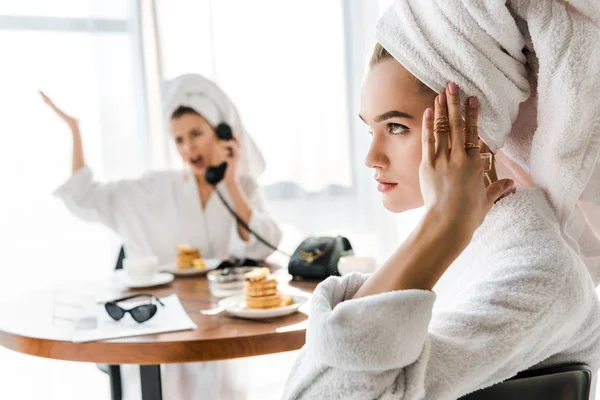 Selective focus of exhausted stylish woman in bathrobe and jewelry with towel on head touching head while emotional friend talking on phone — Stock Image Selective Focus Exhausted Stylish Woman Bathrobe Jewelry Towel Head Touching Stock Picture