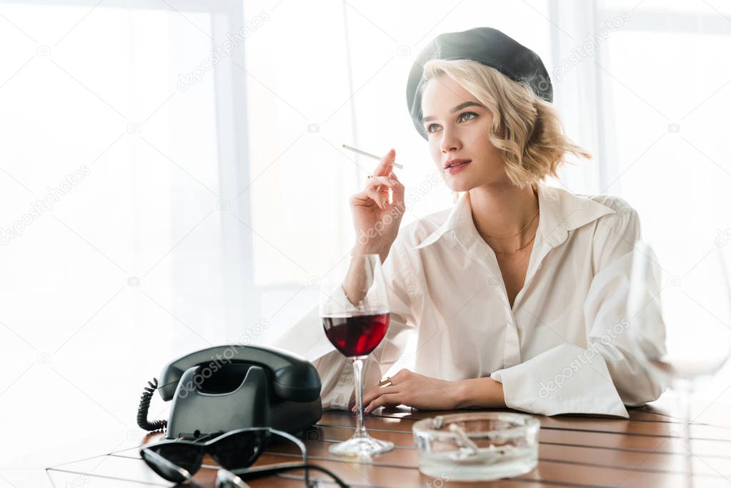 elegant blonde woman in black beret and white shirt smoking cigarette near glass with red wine and vintage telephone