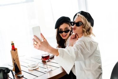 siyah bere ve güneş gözlüğü zarif esmer ve sarışın kadınlar kırmızı şarap içme ve dudakları somurtma ve dil dışarı yapışmasını selfie alarak 