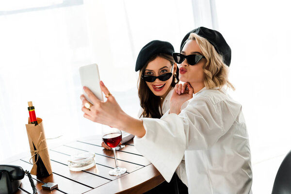 elegant brunette and blonde women in black berets and sunglasses drinking red wine and taking selfie while pouting lips and sticking out tongue 