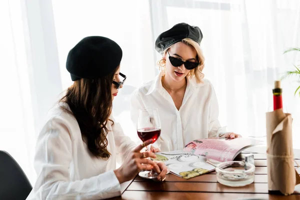 黑发和金发女人在黑色贝雷帽和太阳镜喝红酒和阅读杂志 — 图库照片