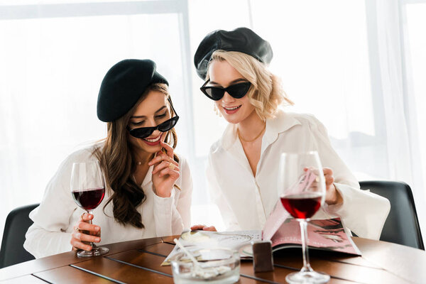 brunette and blonde smiling women in black berets and sunglasses drinking red wine and reading magazine 