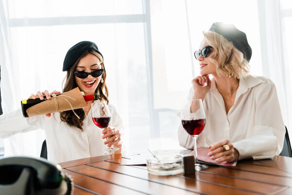 brunette and blonde happy women in black berets and sunglasses pouring red wine