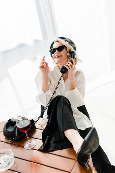 elegant blonde woman in black beret and sunglasses sitting with leg on table, smoking, laughing and talking retro phone