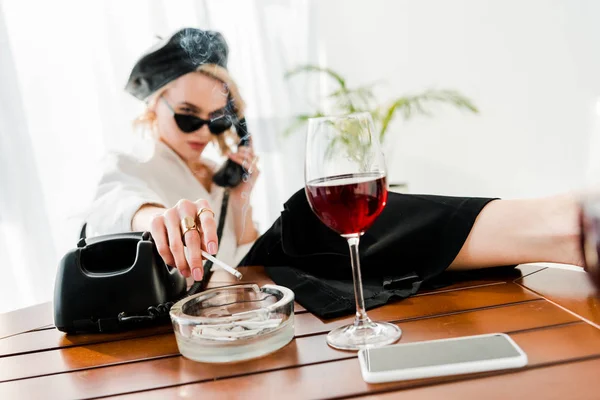 黒いベレーとサングラスで深刻なエレガントなブロンドの女性の選択的な焦点喫煙と赤ワインとガラスの近くにレトロな携帯電話を話す — ストック写真