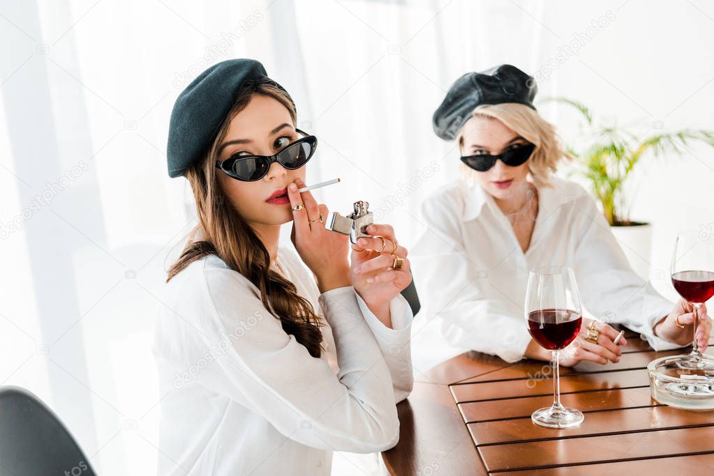 selective focus of elegant woman in black beret and sunglasses lighting up cigarette near friend 