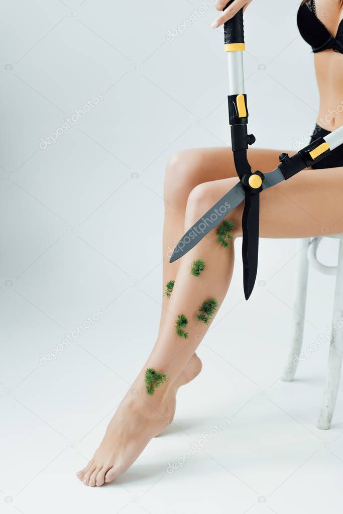 Cropped view of woman sitting on chair and cutting plants on legs with big scissors on grey