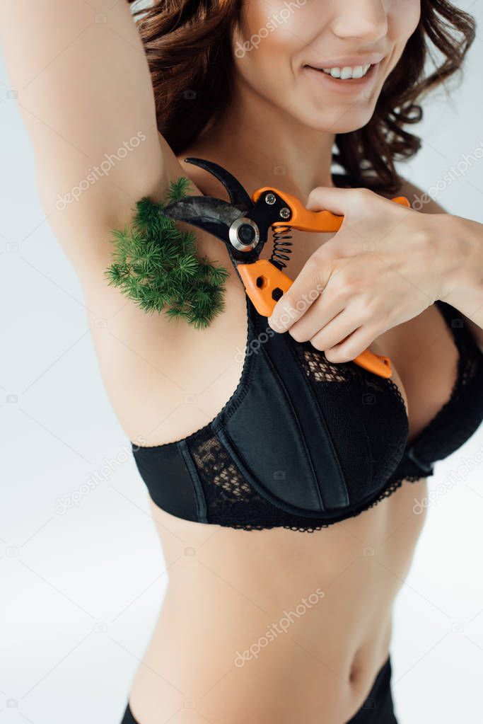 Cropped view of woman in black bra cutting plant on armpit with secateurs on grey
