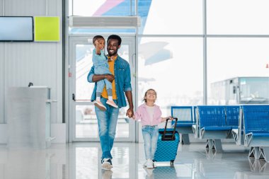 havaalanında bekleyen salon boyunca mutlu çocuklar ve Bagaj ile yürüyüş gülümseyen Afrikalı Amerikalı baba