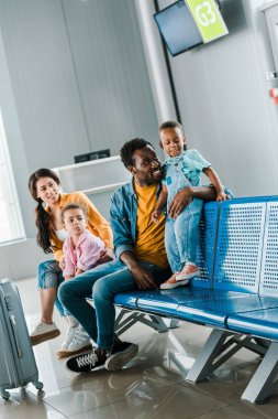 seyahat çantası ve havaalanında oturan çocuklar ile neşeli Afrikalı Amerikan ailesi 