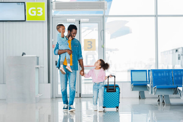 smiling happy african american father walking with children along waiting hall in airport