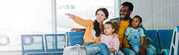 微笑的非洲裔美国家庭与行李和孩子坐在机场的全景镜头 而母亲用手指指着离开 — 图库照片