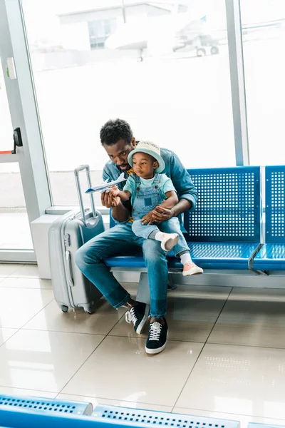 african american father and son sitting with suitcase in waiting hall in airport and playing with toy plane