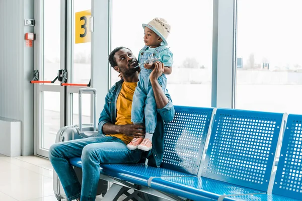 african american father sitting with suitcase in waiting hall in airport and looking at son