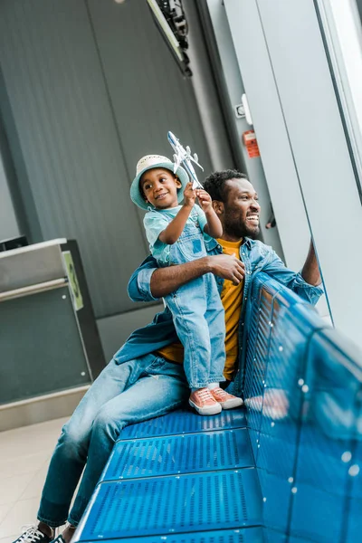 happy african american father looking at window while son playing with toy plane in airport