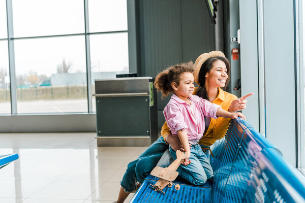 happy african american mother and daughter sitting in airport with wooden plane model  while woman pointing with finger at window