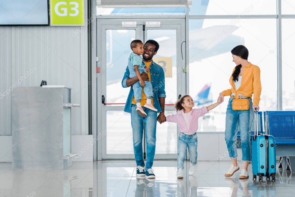 smiling african american family walking with baggage along waiting hall in airport