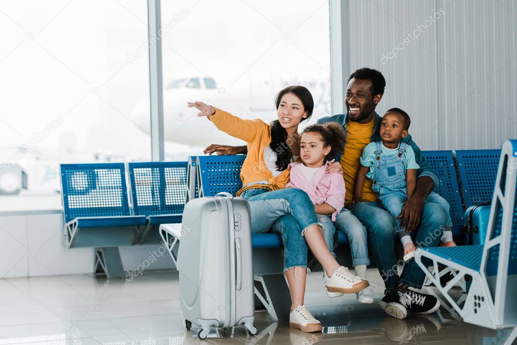 smiling african american family with baggage and kids sitting in airport while mother pointing with finger away