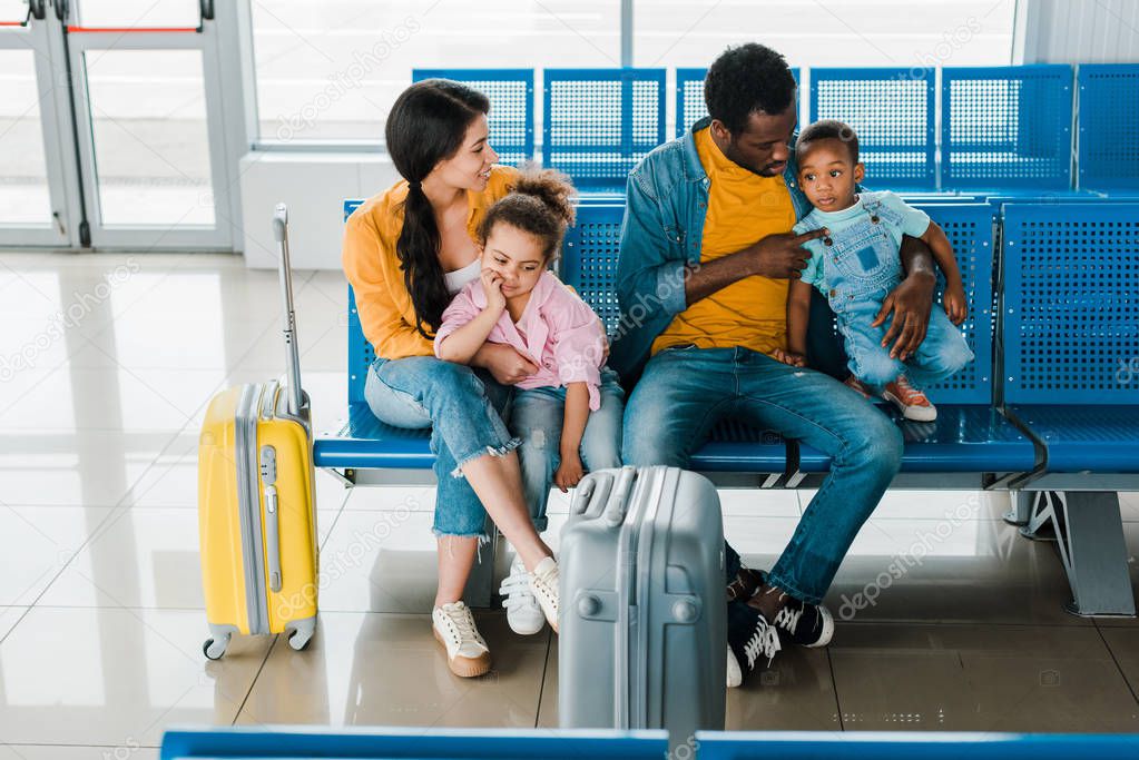 cheerful african american family with travel bags and kids sitting in airport in waiting hall