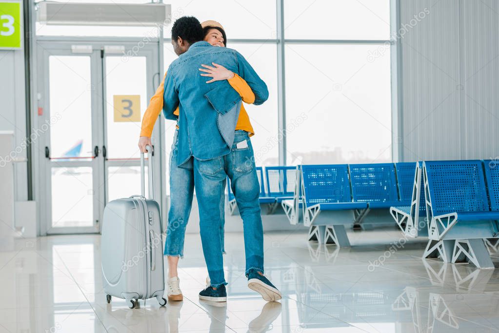 african american man hugging happy girlfriend with suitcase in airport