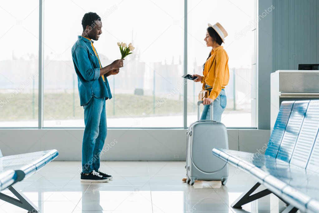 side view of shy african american man holding bouquet of tulips while happy girlfriend walking towards with suitcase in airport