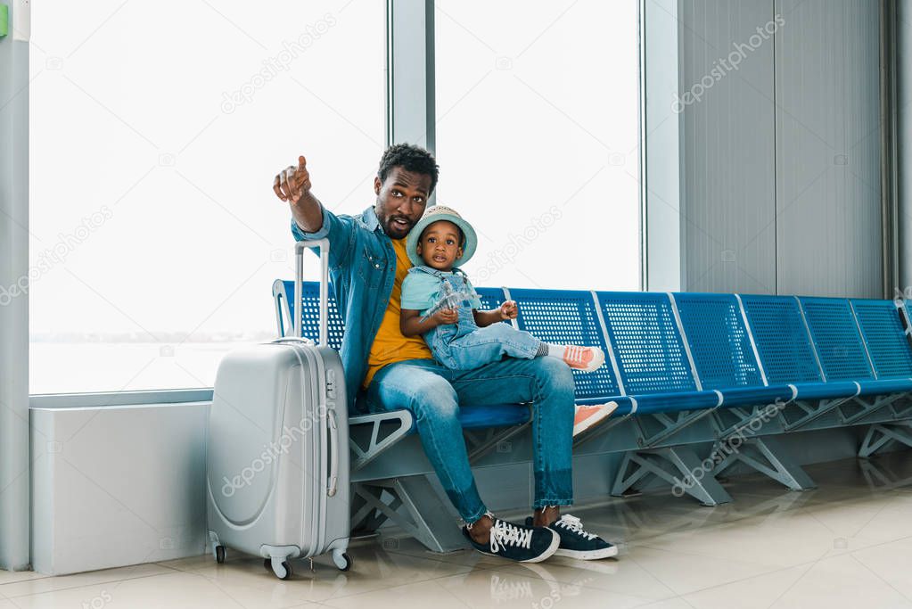 african american father sitting with son in airport and pointing with finger away