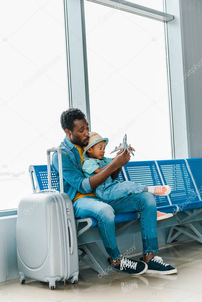 african american father and son sitting with baggage in airport and playing with toy plane