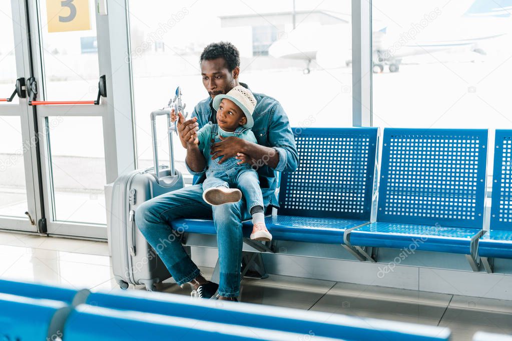 african american father and son sitting in waiting hall in airport and playing with toy plane