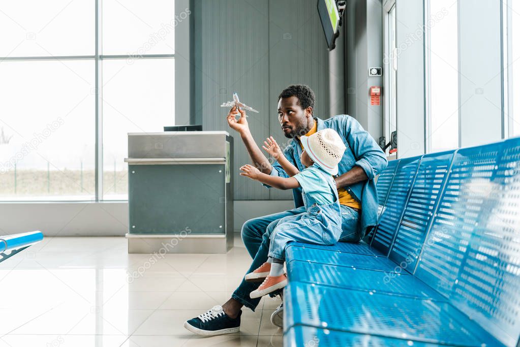 focused african american father and son sitting in waiting hall in airport and playing with toy plane 
