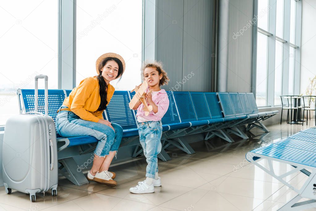 happy african american mother and daughter in airport with suitcase and wooden plane model 
