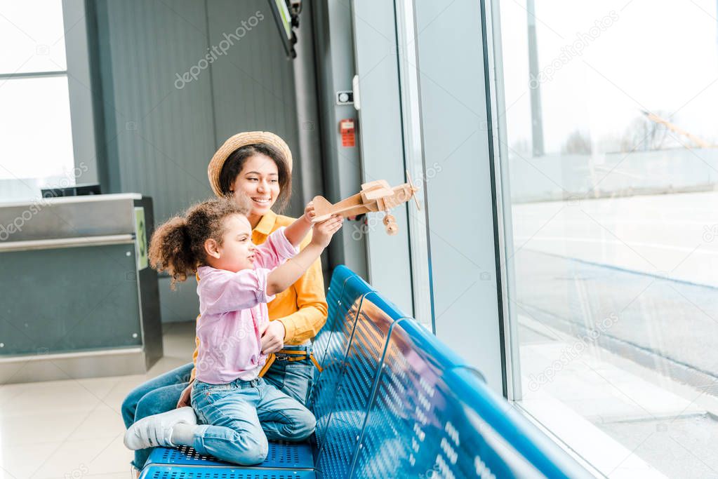 happy african american mother and daughter sitting in airport and playing with wooden plane model