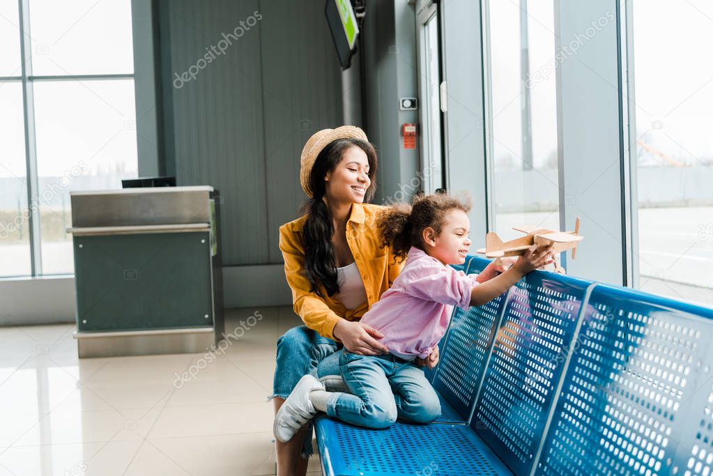 happy african american mother and daughter sitting in airport near window and playing with wooden plane model