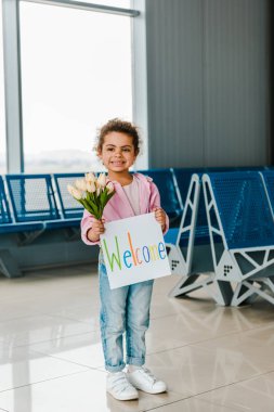 havaalanında bekleyen salonda mutlu Afrikalı Amerikalı çocuk ayakta ve karşılama yazısı ile lale ve plasard tutarak