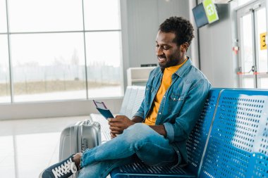 smiling african american man sitting in airport with air ticket and passport clipart