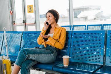 mutlu Afrikalı Amerikan kadın bavul ile kalkış salonunda oturan, kahve gitmek ve akıllı telefonda konuşmak
