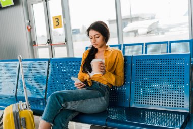 üzgün Afrikalı Amerikan kadın bavul ile kalkış salonunda oturan, kahve gitmek ve akıllı telefon kullanarak