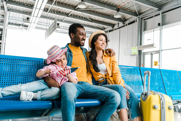 smiling african american family sitting in airport with suitcase while kid holding american flag
