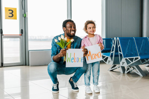 african american daughter and father standing in waiting hall in airport and holding tulips and placards with welcome mama words
