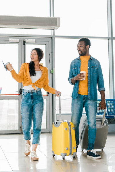 happy african american couple walking together in airport with travel bags and coffee to go